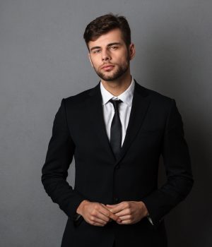 Close-up photo of young successful business man in black suit looking at camera, isolated on gray background